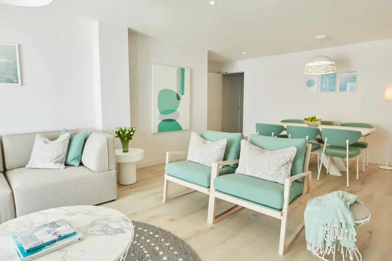 Modern luxury hotel living room with teal chairs, white sofa, dining table, and abstract art.