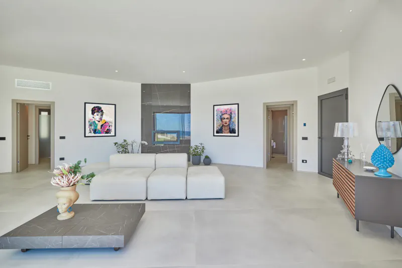 Spacious modern luxury living room with white modular sofas, gray coffee table, colorful art prints, plants, and sea view fireplace.