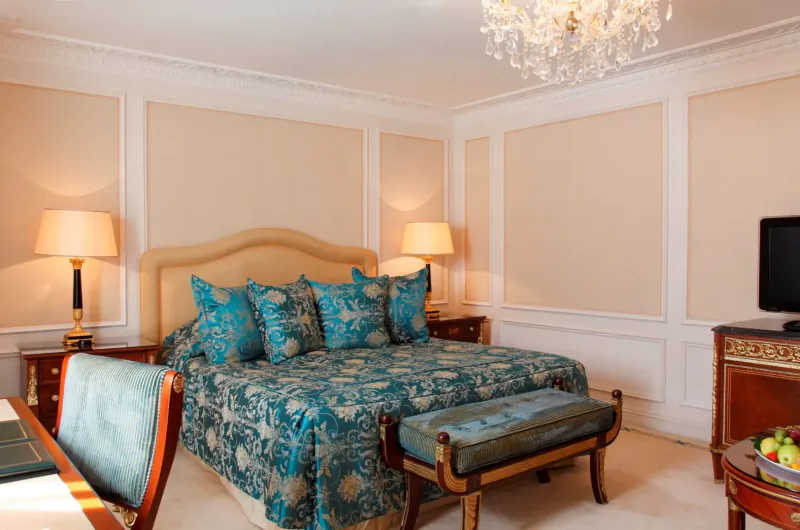 Elegant Bentley Hotel bedroom with turquoise bedspread, chandelier, lamps, TV, bench, and fruit bowl.