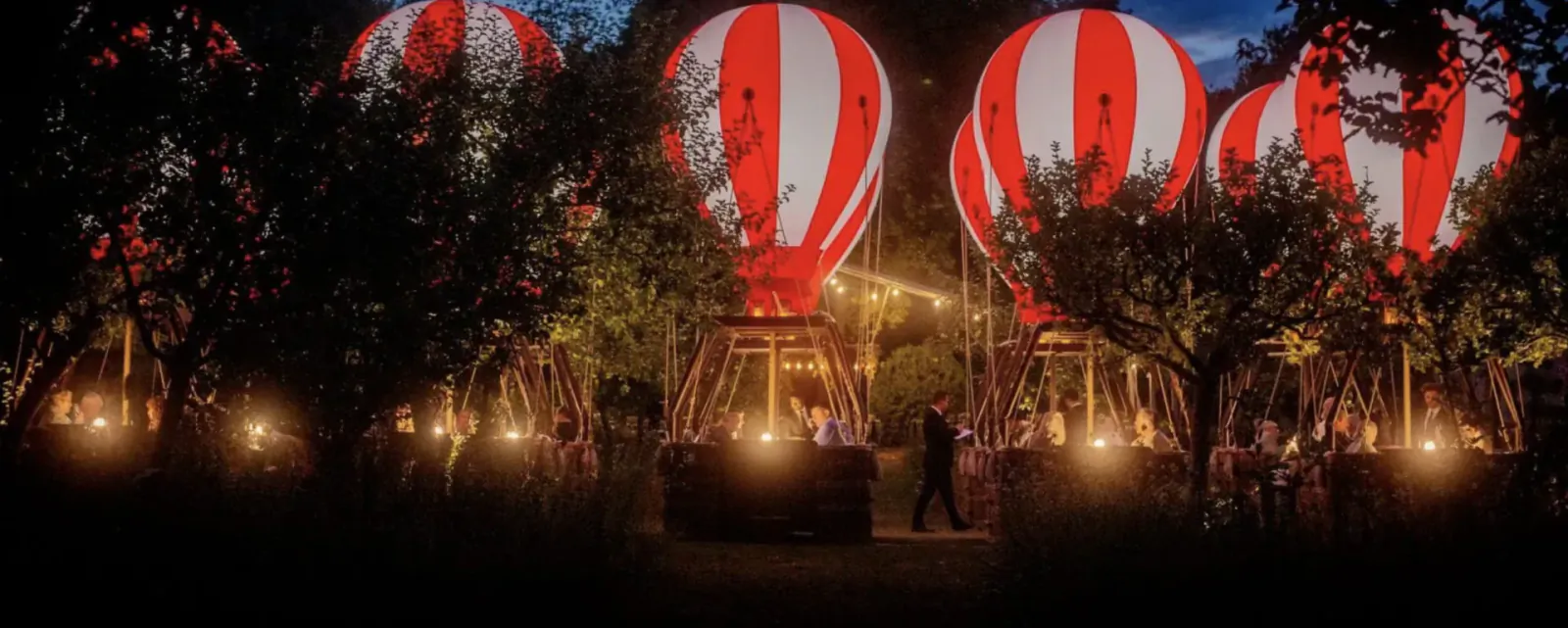 Illuminated red and white hot air balloons glowing among trees at night in The Grove London's country retreat, with a person walking nearby.