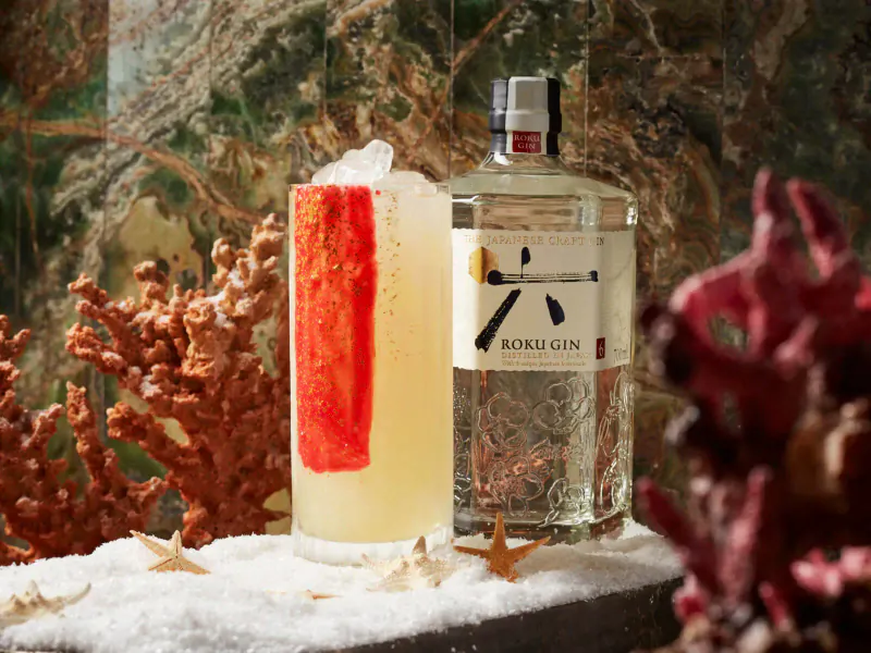 Roku Gin bottle beside tall glass of yellow cocktail with red rind garnish, set against coral and seaweed backdrop.