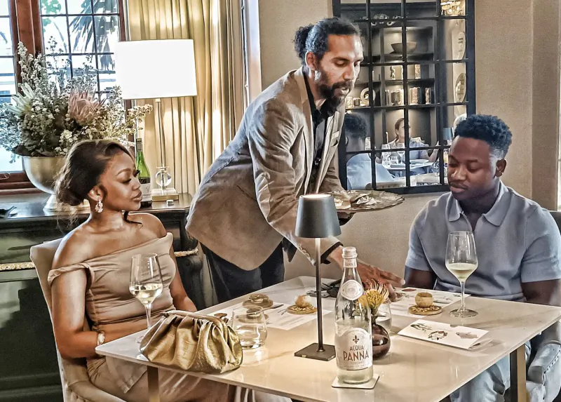 Elegant trio at candlelit dinner table: man in suit serves wine to woman in off-shoulder dress and man in polo, De Tafel bottle visible.
