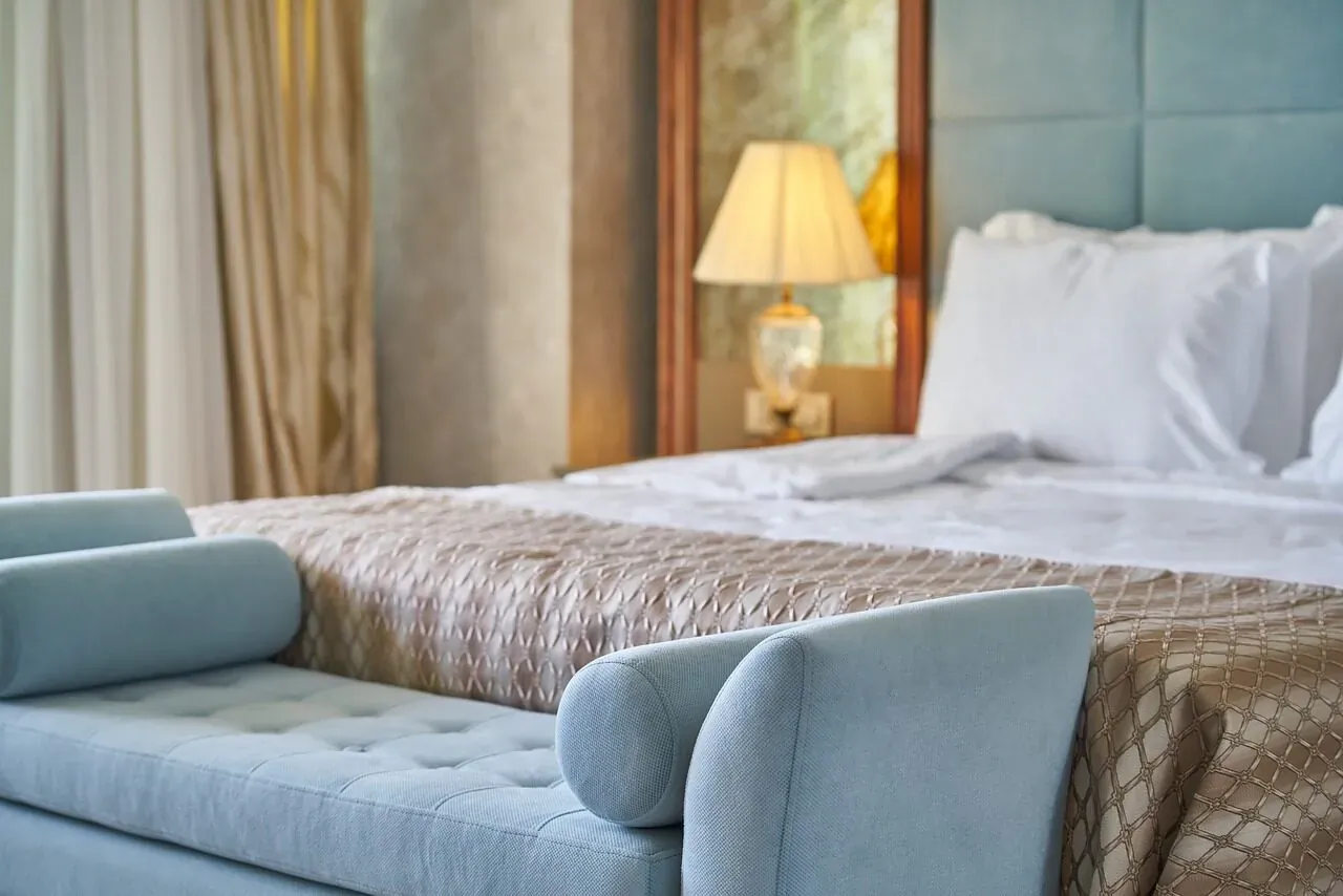 Luxury hotel bedroom with plush bed, turquoise bolster pillows, beige throw, lamp, and curtains