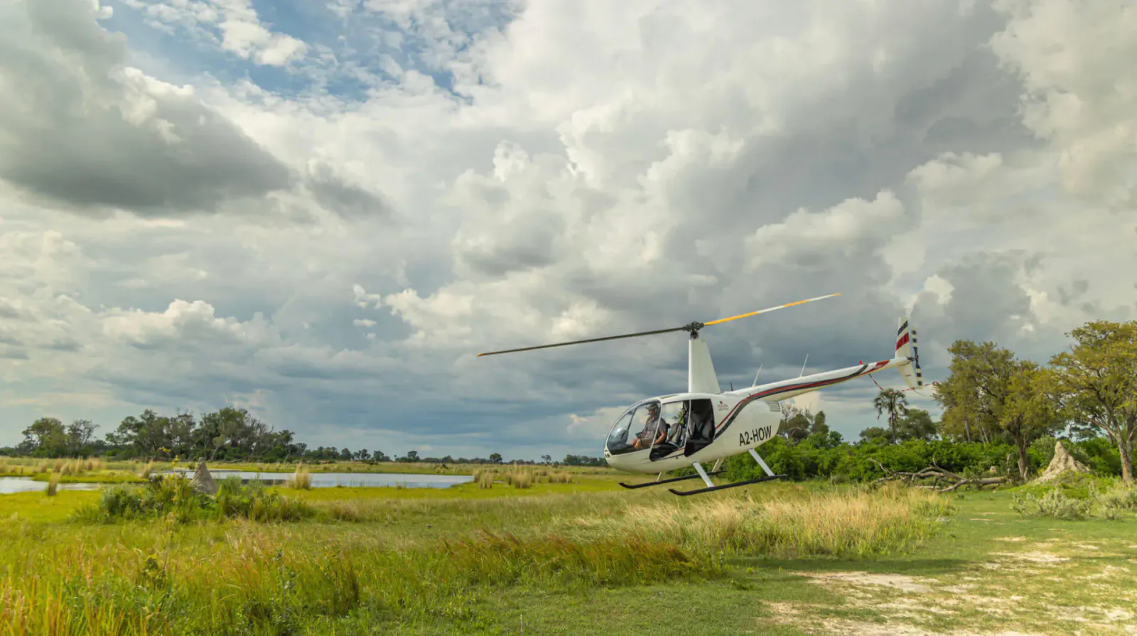 White Mauna Delta Helicopter with yellow tail rotor on grassy landing area by lake under cloudy sky, pilot inside.