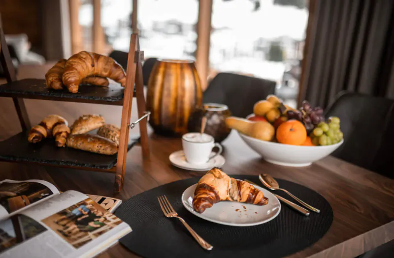 Luxury chalet breakfast table with croissants on tiered stand, coffee cup, fruit bowl of oranges and grapes, magazine nearby.