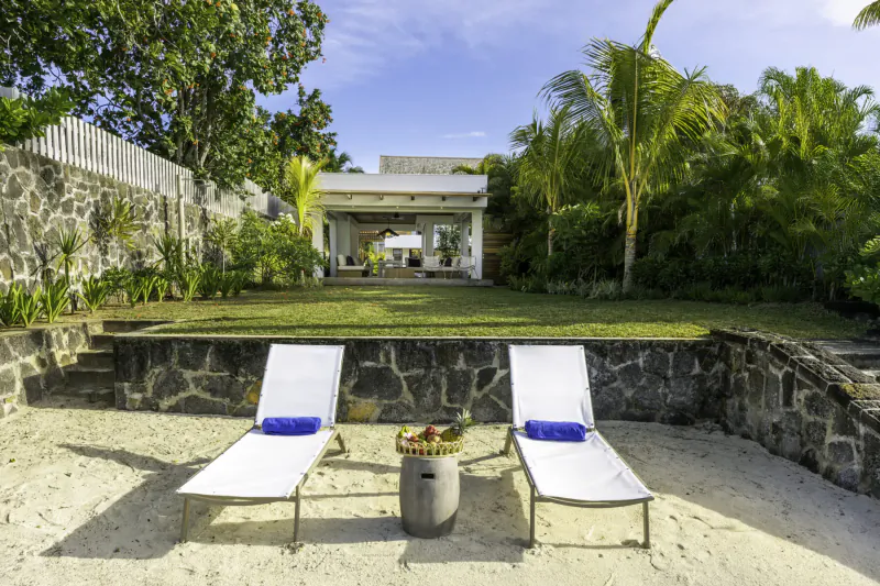 Two white lounge chairs with blue towels and fruit basket on Belle Mare beach, Villa Diamond with lush palms behind stone wall.