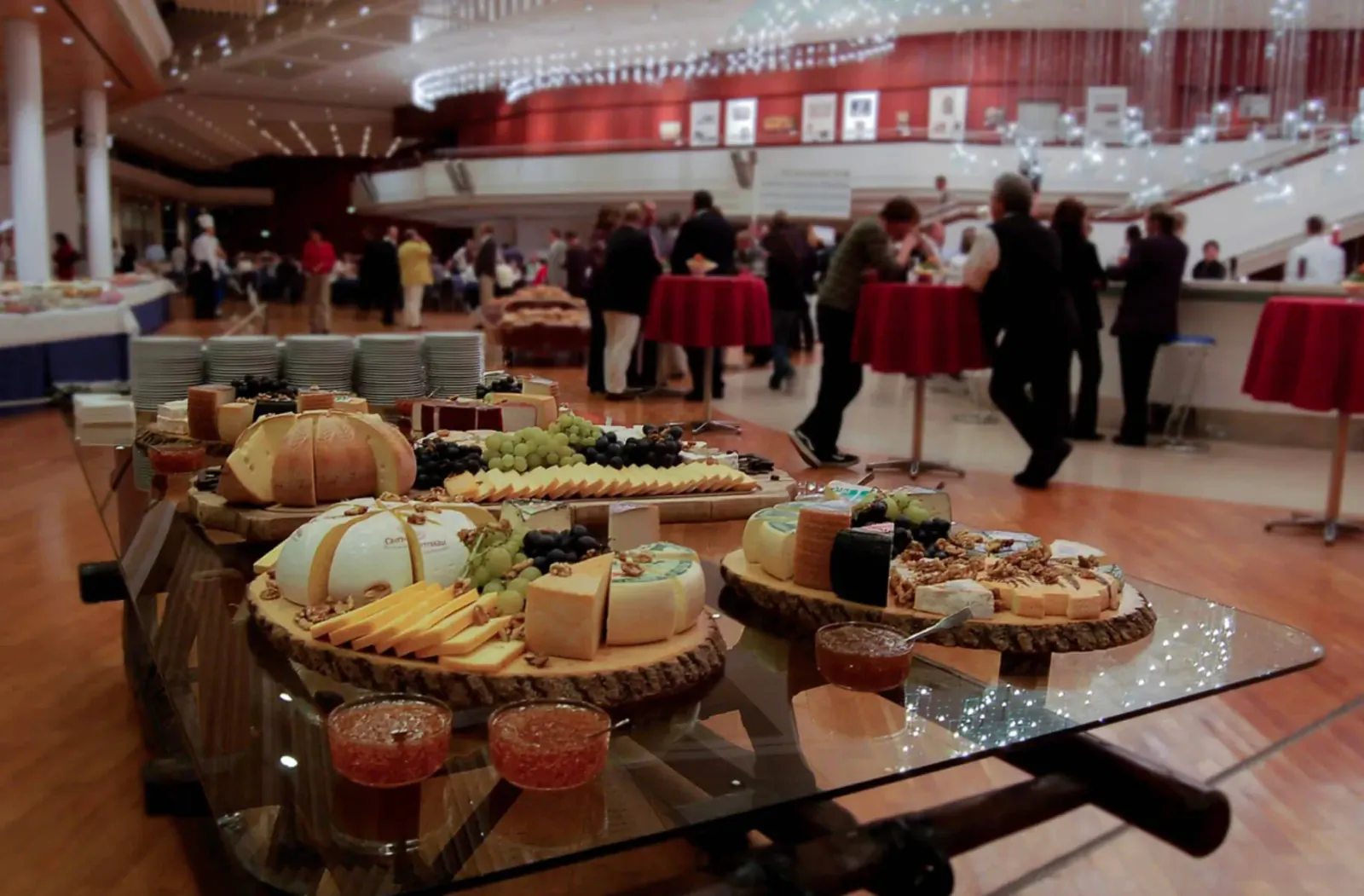 Assortment of cheeses, breads, fruits on wooden boards on glass table at elegant event venue with chandeliers and crowds.