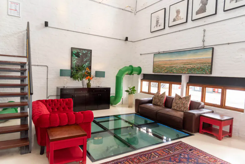 Luxury hotel gallery lounge with red tufted sofa, green slide, glass floor, eclectic art, and large windows.