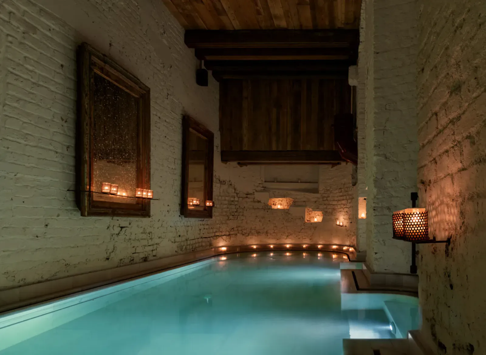 AIRE Ancient Baths: candlelit indoor pool in rustic white brick room with wooden beams and mirrors