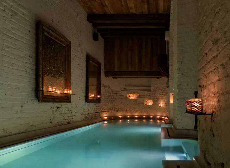 AIRE Ancient Baths: candlelit indoor pool in rustic white brick room with wooden beams and mirrors
