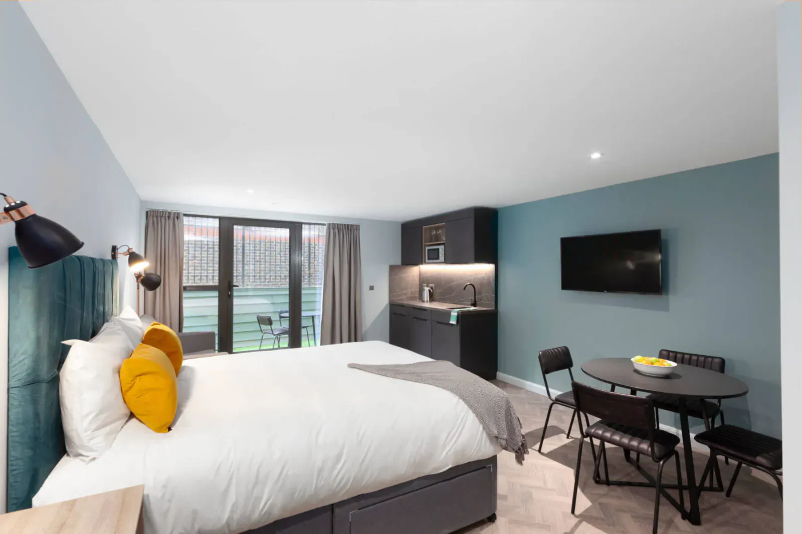 Modern aparthotel room with king bed, yellow pillows, kitchenette, dining table, TV, and balcony view.
