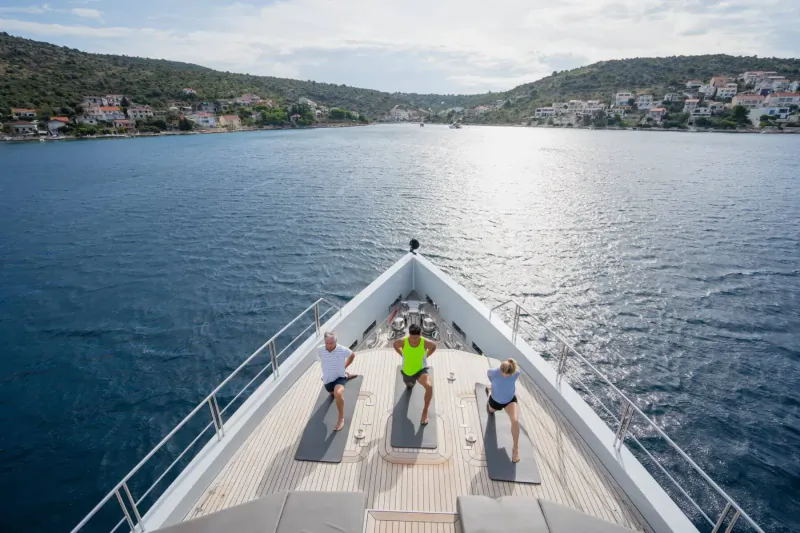 Three people doing yoga poses on the bow of a luxury yacht overlooking a scenic coastal bay.