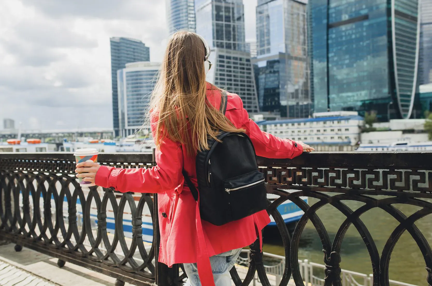 Woman in red coat and backpack holds coffee cup, leaning on railing overlooking Moscow River and skyscrapers