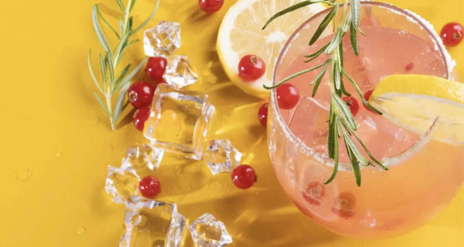 Pink cocktail garnished with rosemary sprigs, redcurrants, lemon slices, and ice cubes on yellow background