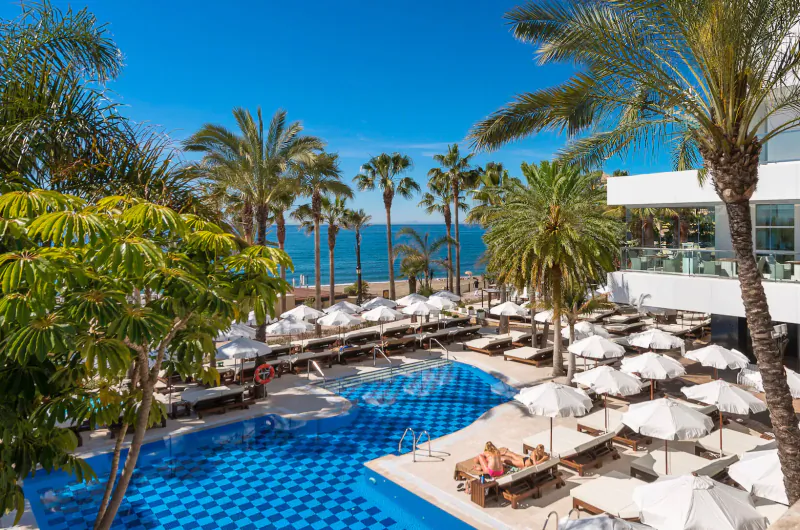Aerial view of Amàre Marbella's blue mosaic pool with loungers, white umbrellas, palm trees, and sea beyond under blue sky.