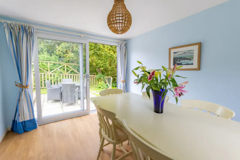 Light blue dining room with white table, pink lilies in purple vase, rattan lamp, and sliding doors to garden patio.