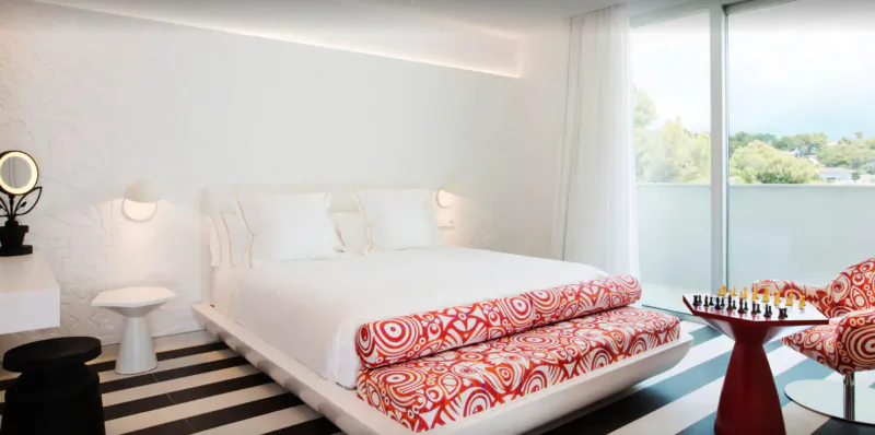 Marcel Wanders-designed hotel bedroom with white bed, orange patterned bench, chess set, ocean-view balcony.