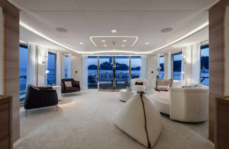 Luxurious yacht interior with white leather seating, ottomans, and panoramic windows overlooking dusk sea.