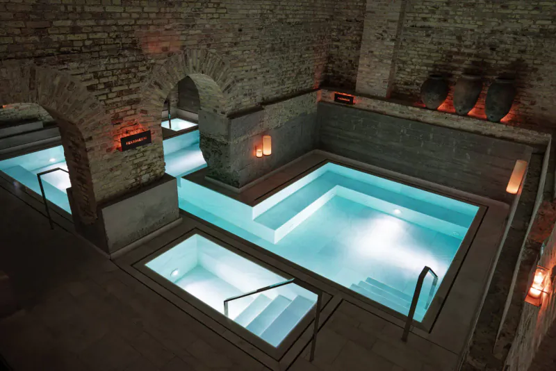 Aerial view of candle-lit luxury pools in brick arched ancient baths, glowing turquoise water and steps.