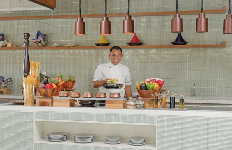 Chef in white uniform holding a plate of colorful fruits in luxurious Maldives resort kitchen with copper lamps and fresh produce