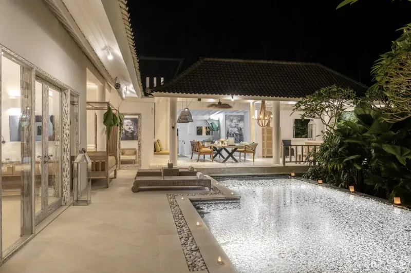 Luxurious Bali villa at night with infinity pool, dining table, lounge area, and tropical plants