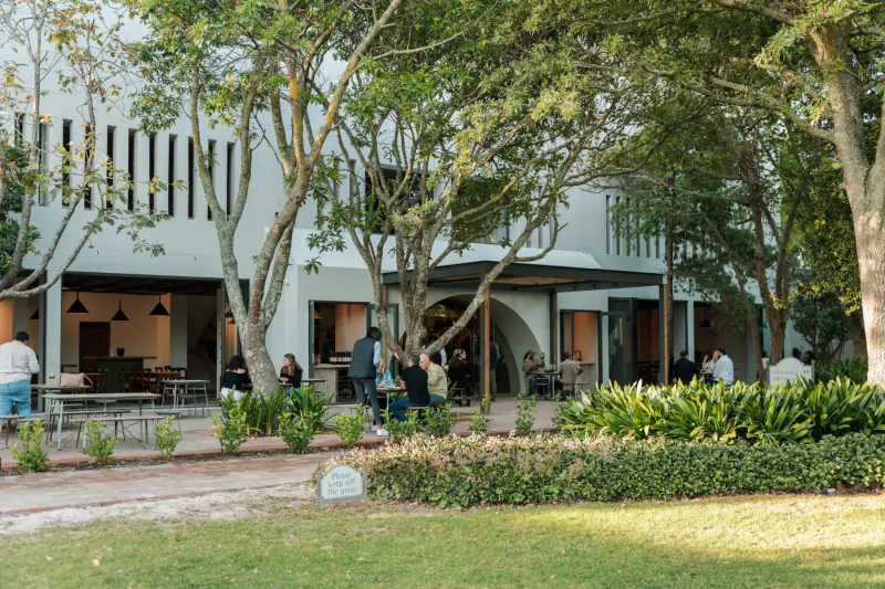 Modern white gallery at Groot Constantia with arched entrance, trees, patrons at outdoor tables, lush gardens.