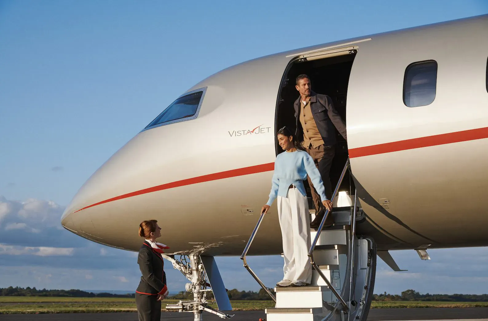 Flight attendant assists stylish couple boarding VistaJet private jet on tarmac under blue sky