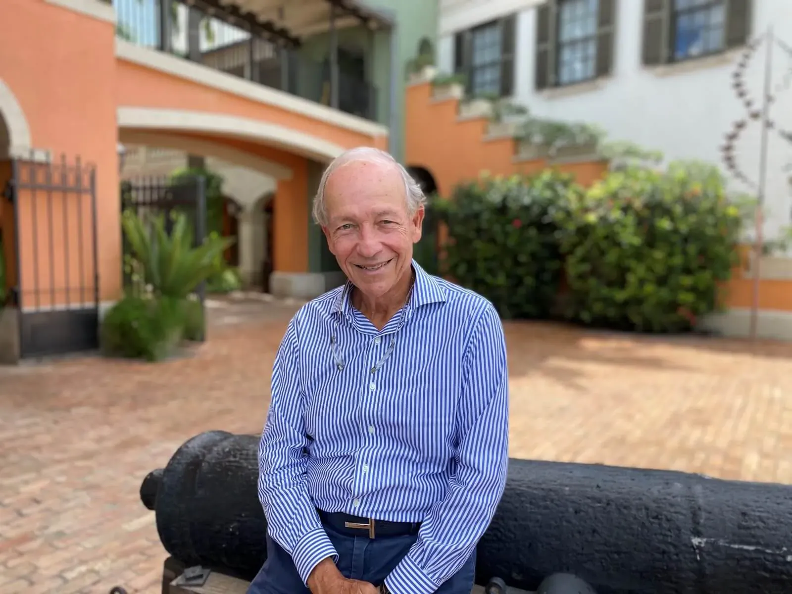 Sir Paul Altman smiling, seated on a black cannon in a colorful Caribbean courtyard with orange buildings and greenery.