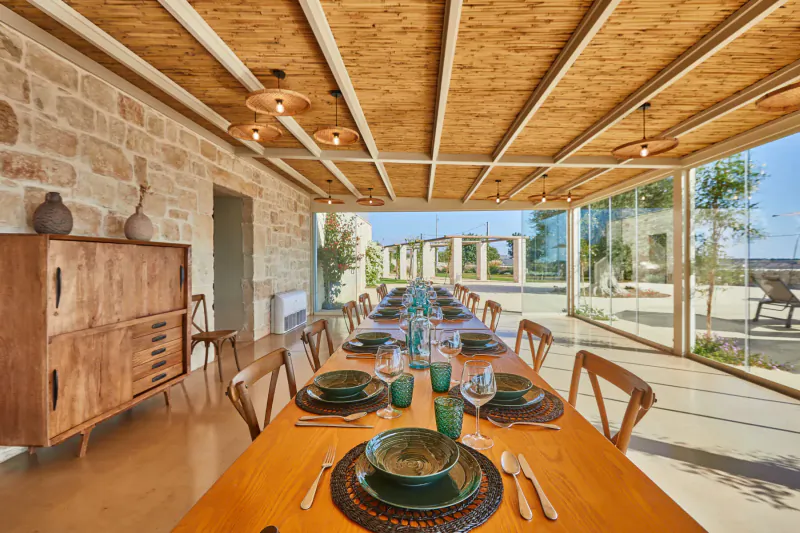 Elegant rustic dining room with long wooden table set for meal, green plates, glass walls, stone walls, and garden view in Sicily hotel.