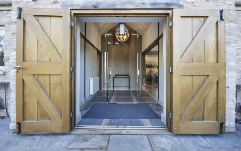 Open wooden barn doors framing entrance to luxury cottage hallway with chandelier and stone walls
