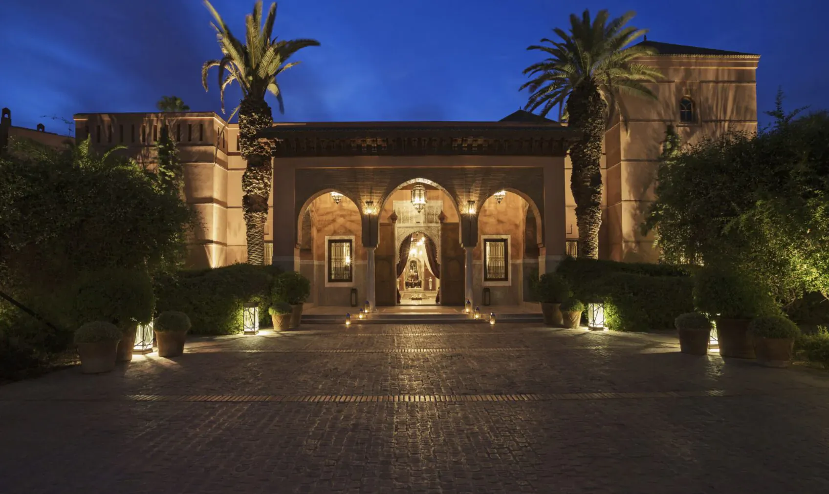 Night view of Royal Mansour Marrakech's grand arched entrance, lit warmly with palm trees and lanterns in courtyard.