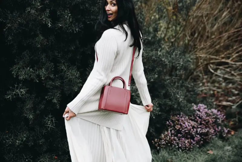 Woman in white pleated dress and cardigan with red Luxtra handbag, smiling over shoulder amid green shrubs