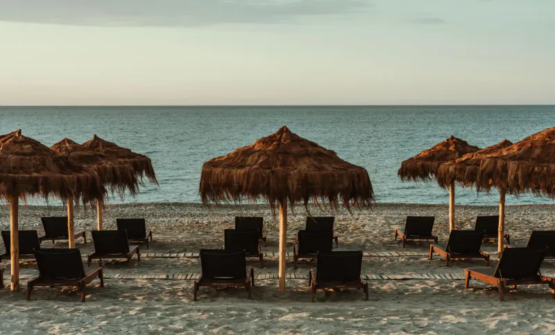 Row of thatched beach umbrellas shading black lounge chairs on sandy shore by calm sea at sunset, Asterion Suites & Spa, Crete