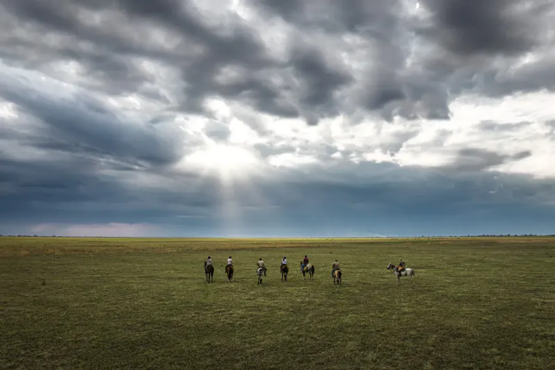 Silhouettes of horseback riders on vast green savanna under dramatic stormy sky with sun rays, Zambian safari.