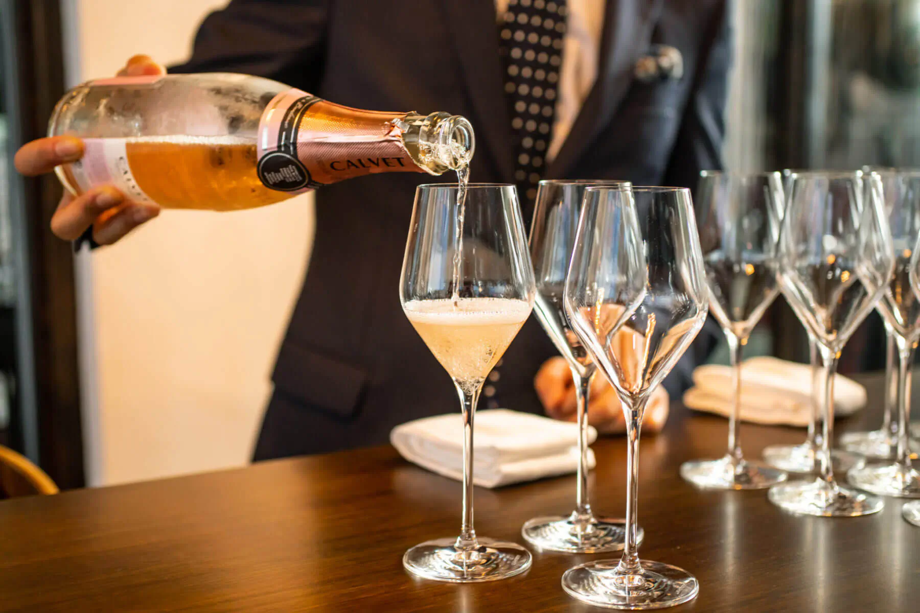 Man in suit pouring champagne from bottle into rows of tall glasses on wooden table for festive season