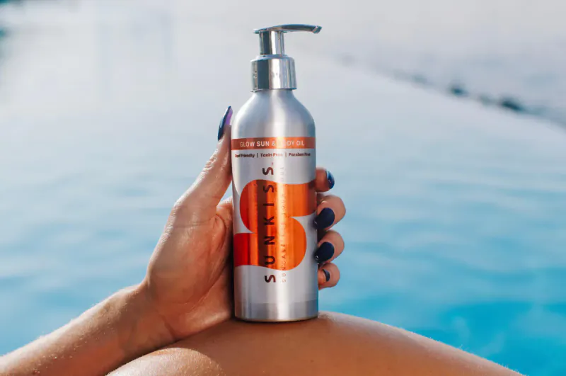 Woman's hand holding silver SunKiss suncare bottle with orange VS logo by blue pool.