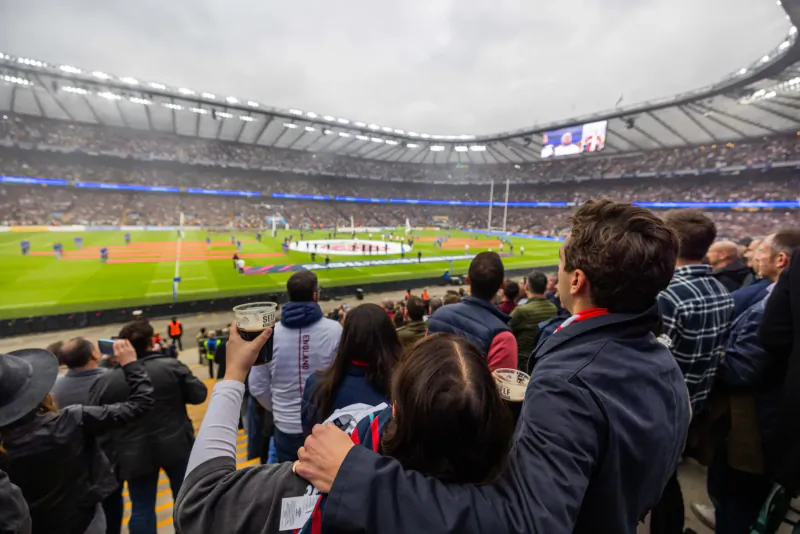 Crowd in stands at packed stadium watching rugby match on field, some holding drinks and phones, cloudy sky.