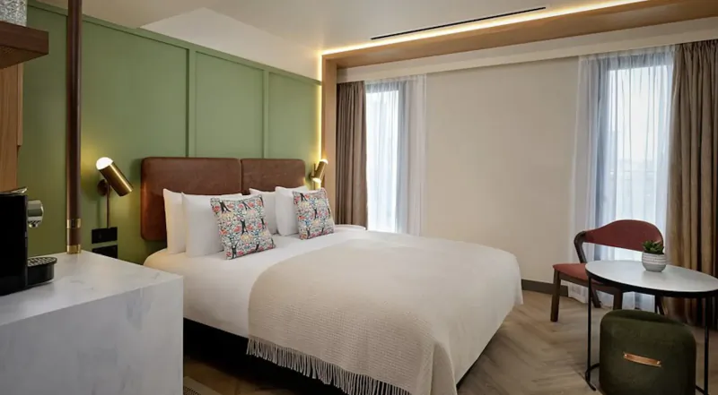 Modern hotel room at The Reach at Piccadilly, Manchester, with king bed, green accent wall, Nespresso machine, chair, and window views.
