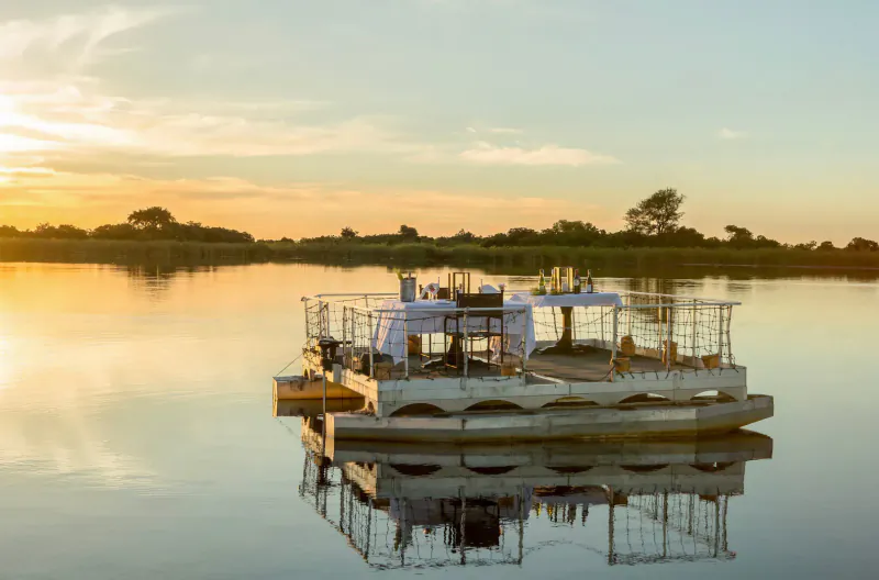 White floating lodge on calm lagoon at sunset, with tables, chairs, and reflection in golden light, Xugana Island Lodge