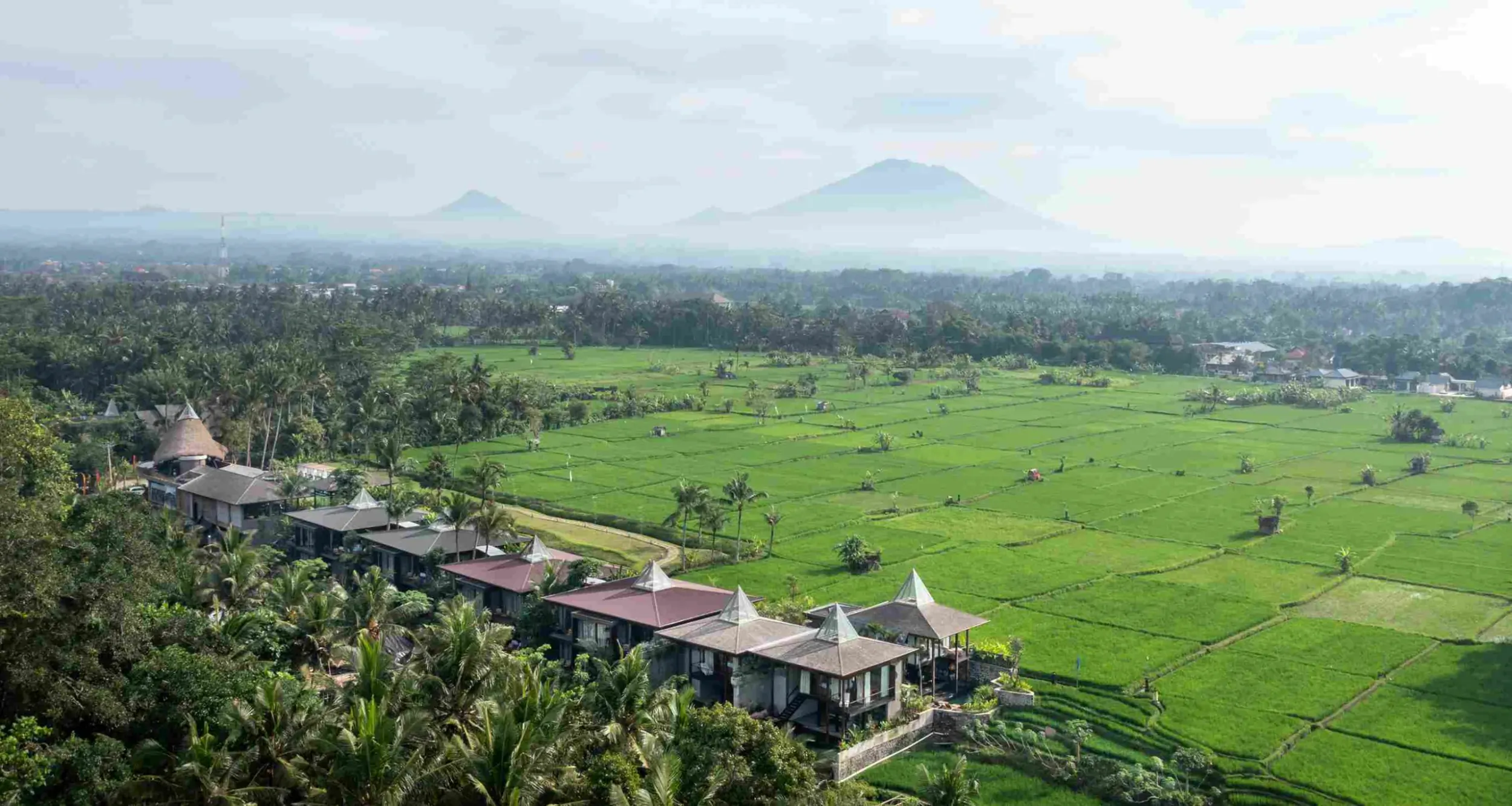 Aerial view of GDAS Bali luxury retreat with thatched villas amid green rice fields and distant volcanoes.