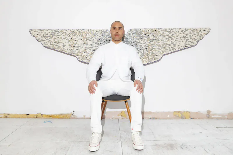 Bald man in white suit sits on wooden chair with large stone angel wings on white wall behind him at Aston Martin Residences art gallery