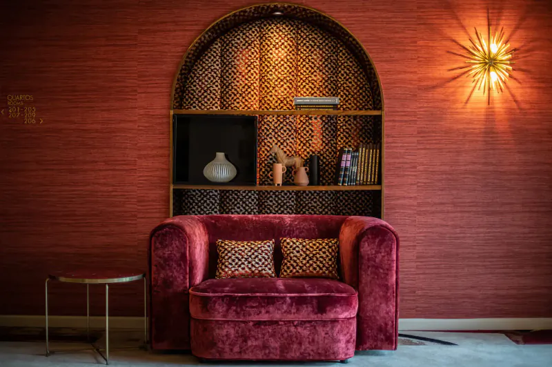 Cozy deep pink velvet armchair with pillows in luxurious red alcove shelving with vase, books, and starburst wall lamp at VINHA Boutique Hotel