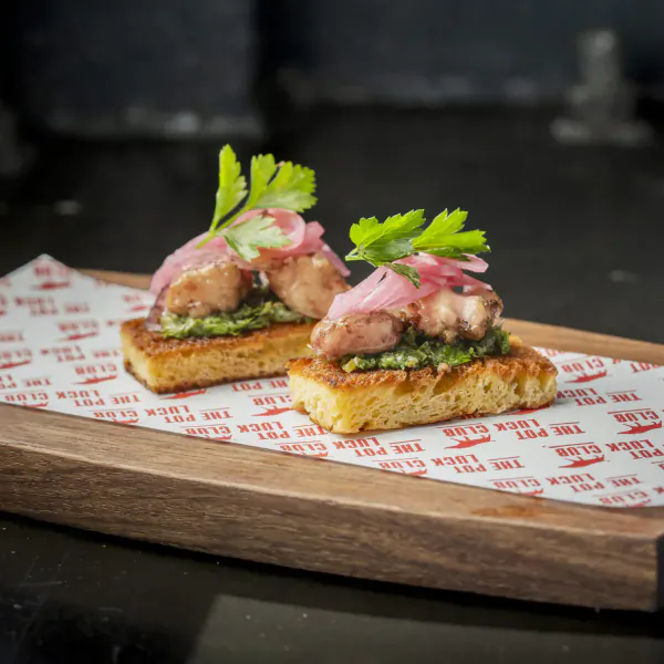Two grilled chicken skewers on chickpea pancakes with green chutney, pink onions, and cilantro on a wooden board
