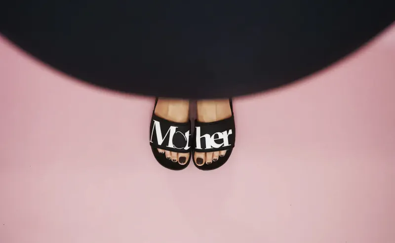 Overhead view of black flip-flops with 'MoTHER' text on pink background, toes visible.