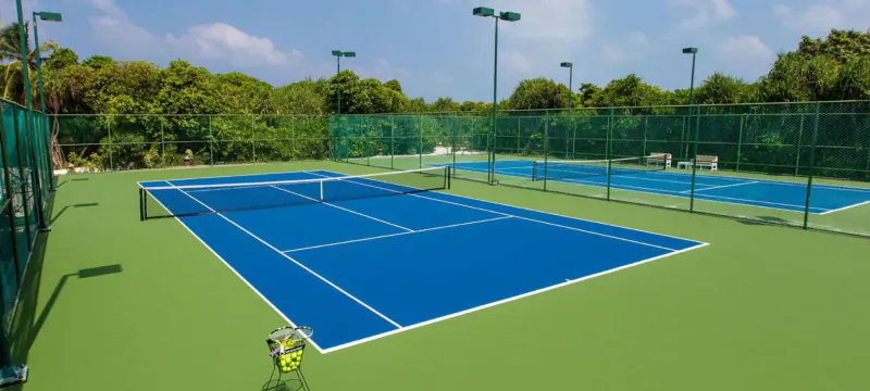 Two blue tennis courts with green padding, surrounded by green mesh fences and lush trees under a clear sky