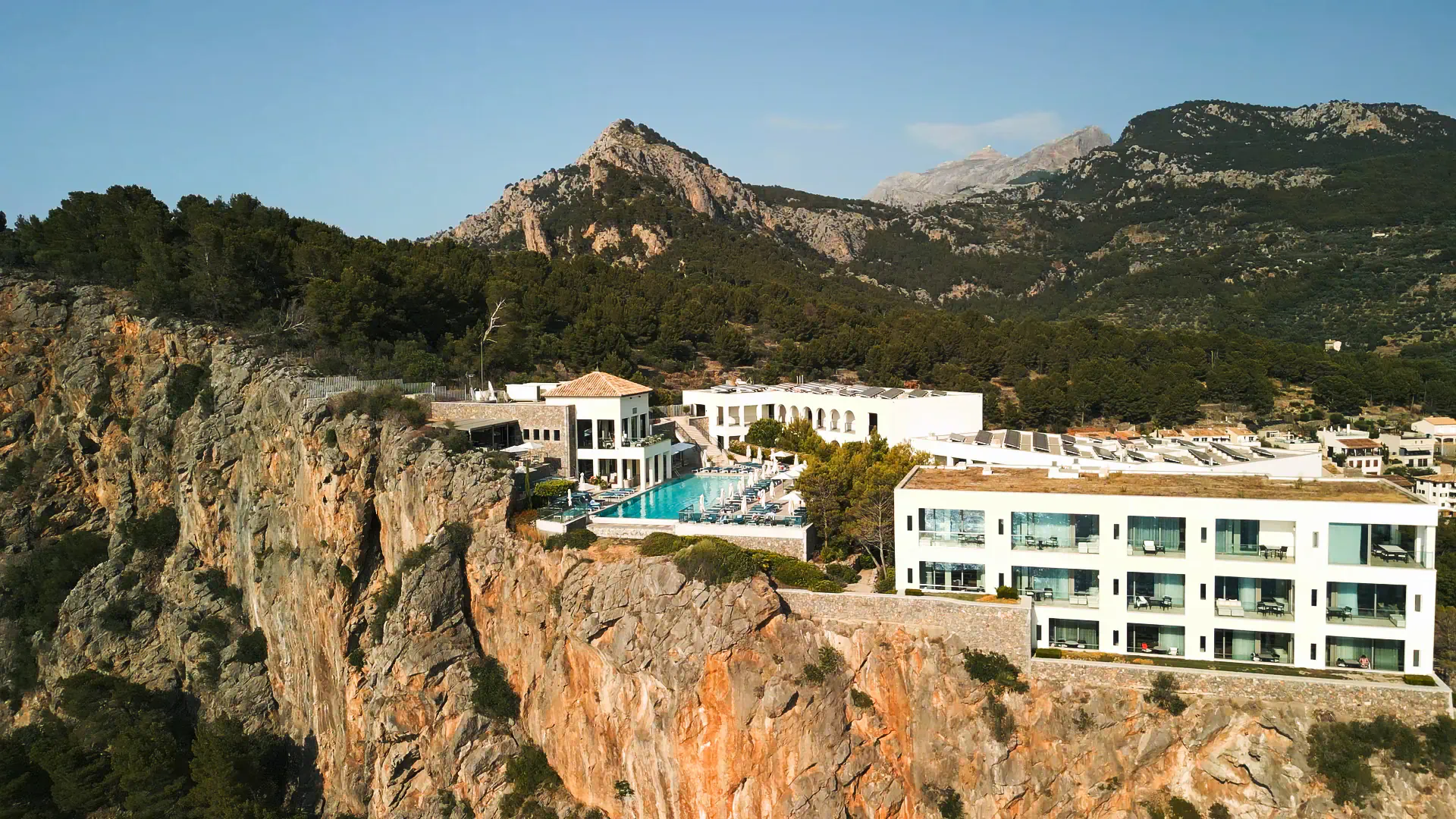 Aerial view of Jumeirah Mallorca resort with infinity pool on cliffside overlooking mountains and sea.