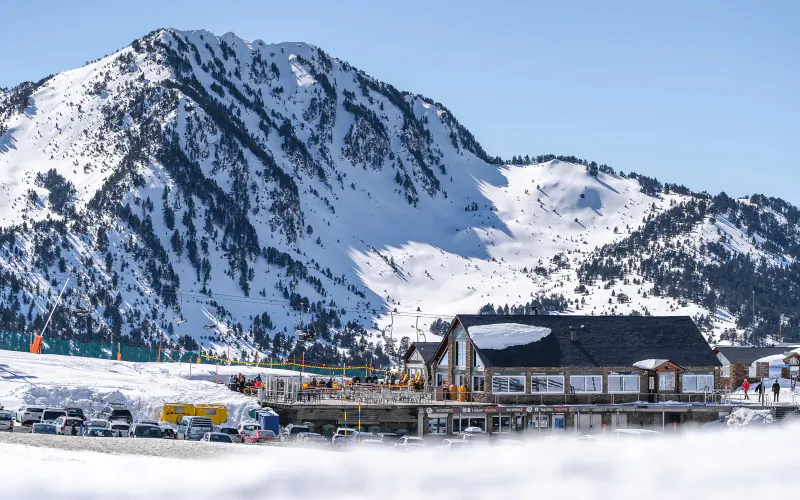 Snowy Pyrenees mountains in Val d'Aran with wooden ski lodge, vehicles, and skiers under blue sky
