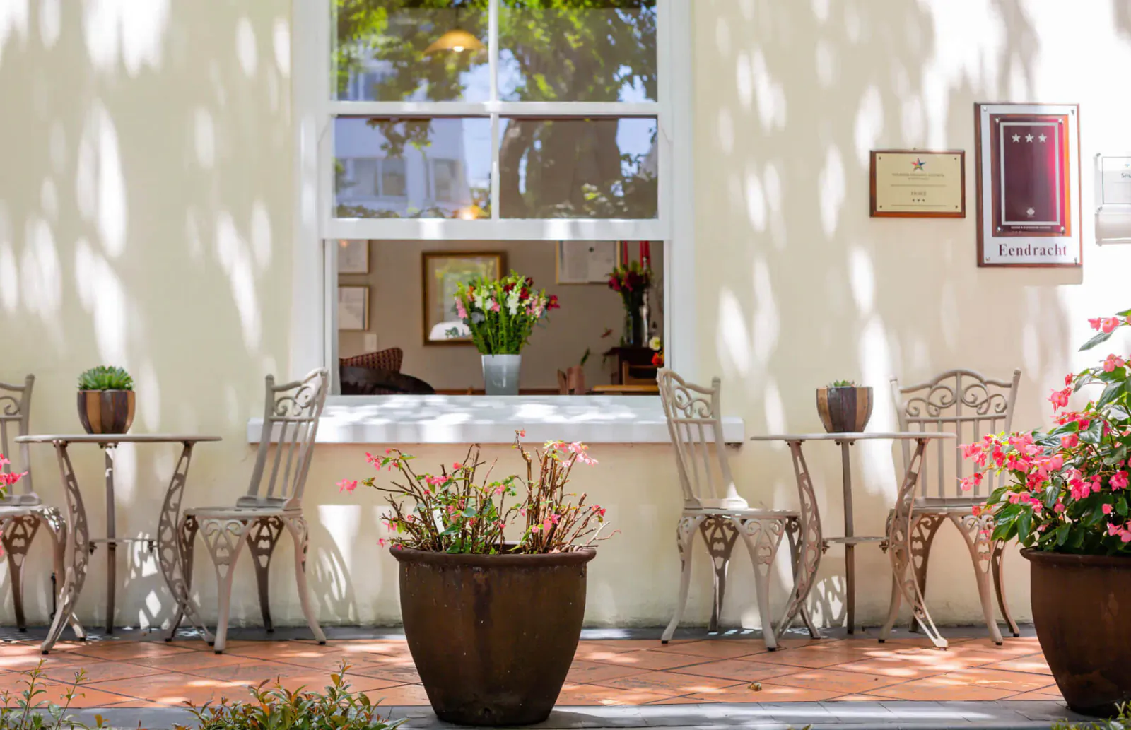 Outdoor café at Eendracht Hotel with ornate metal tables, chairs, potted flowers, and window view of interior bouquet.