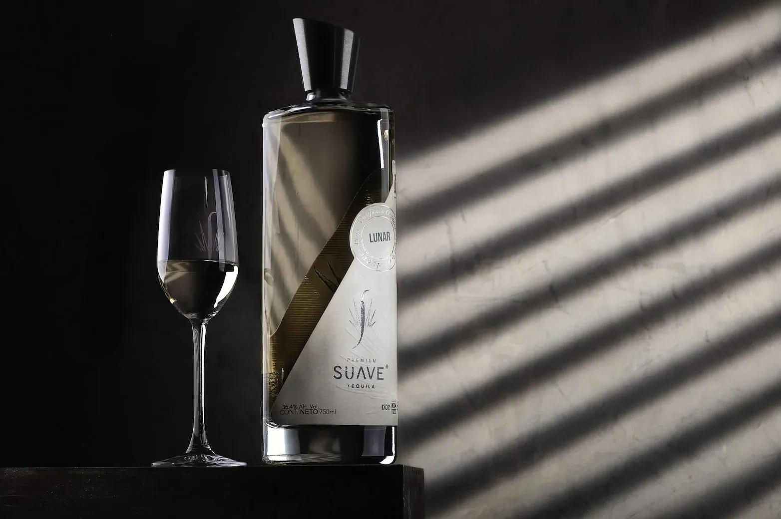 SUAVE Tequila bottle and glass of pale liquor on dark surface with sun rays, for Tequila Day.