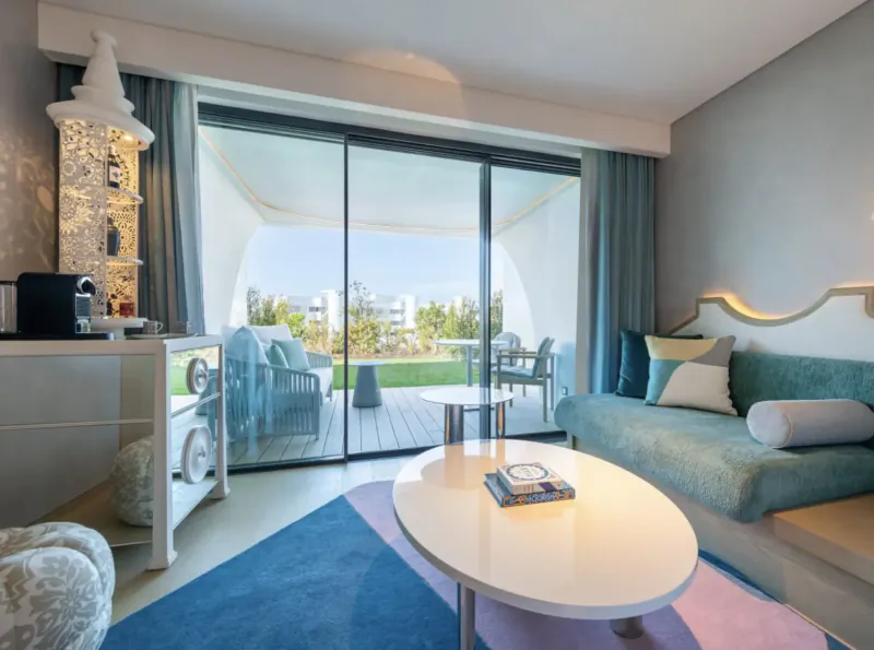 Modern W Algarve hotel suite with teal sofa, coffee table, Nespresso machine, and balcony view of pool and white buildings.